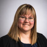 Headshot photo of smiling woman with glasses and brown shoulder length straight hair.